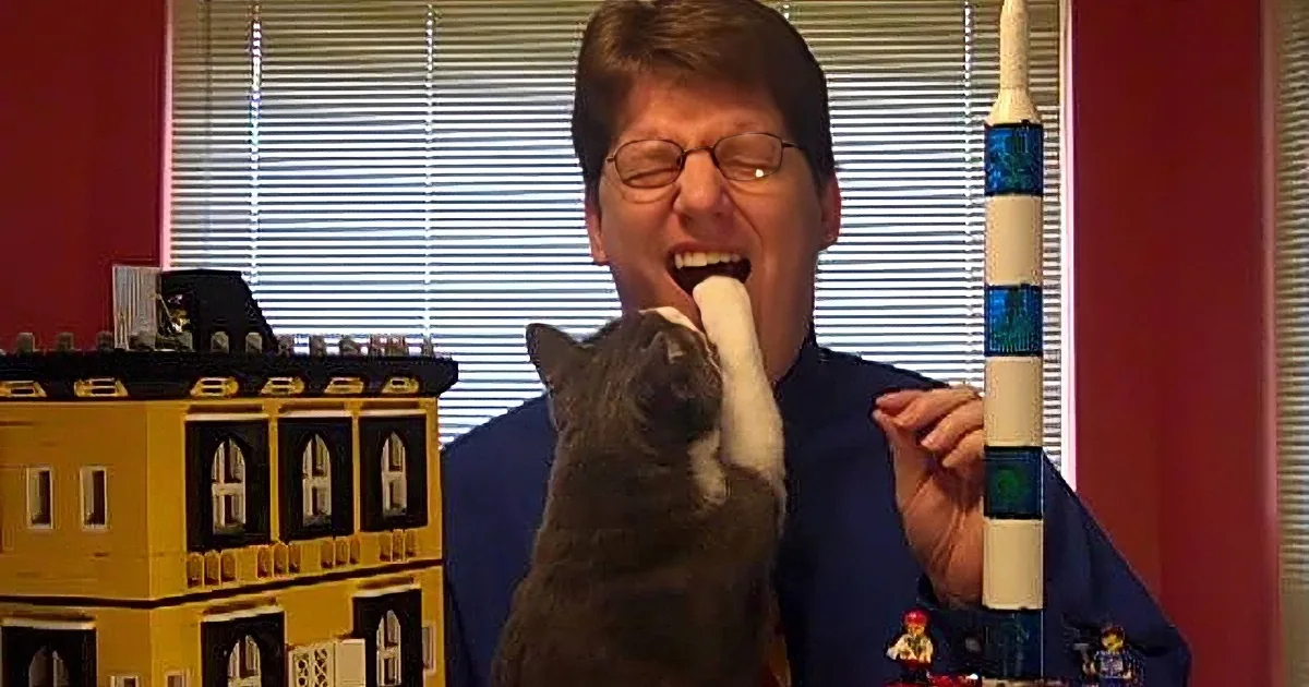 A man in glasses holds a cat while making a funny face, surrounded by colorful Lego-like structures.