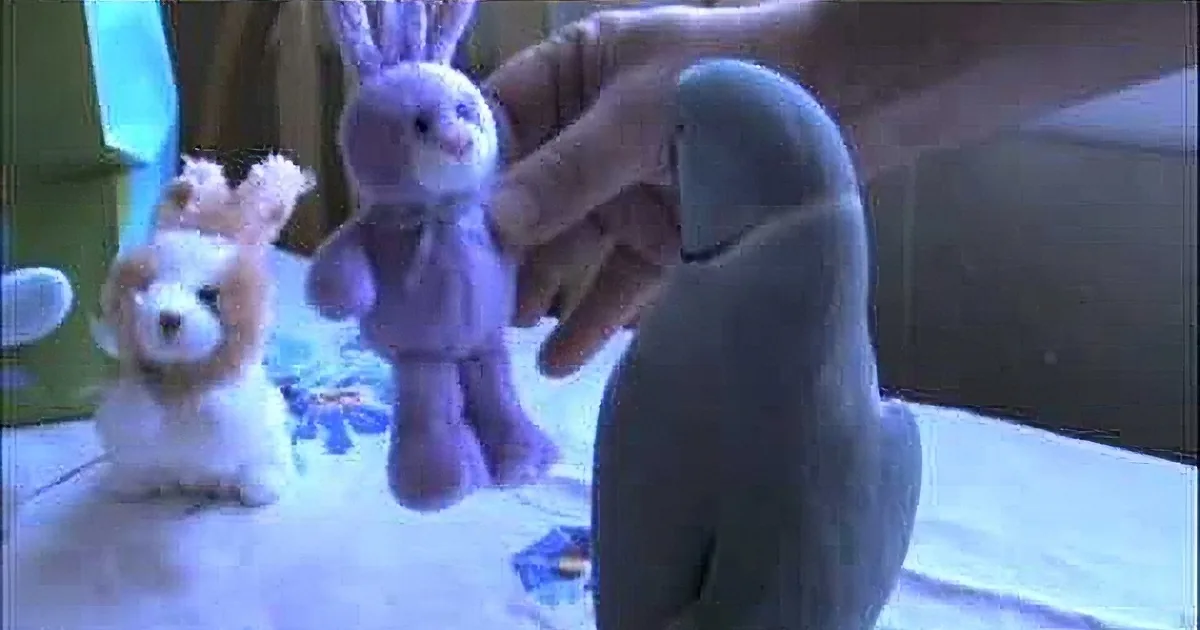 A hand holds a purple stuffed rabbit, surrounded by other plush toys on a table.