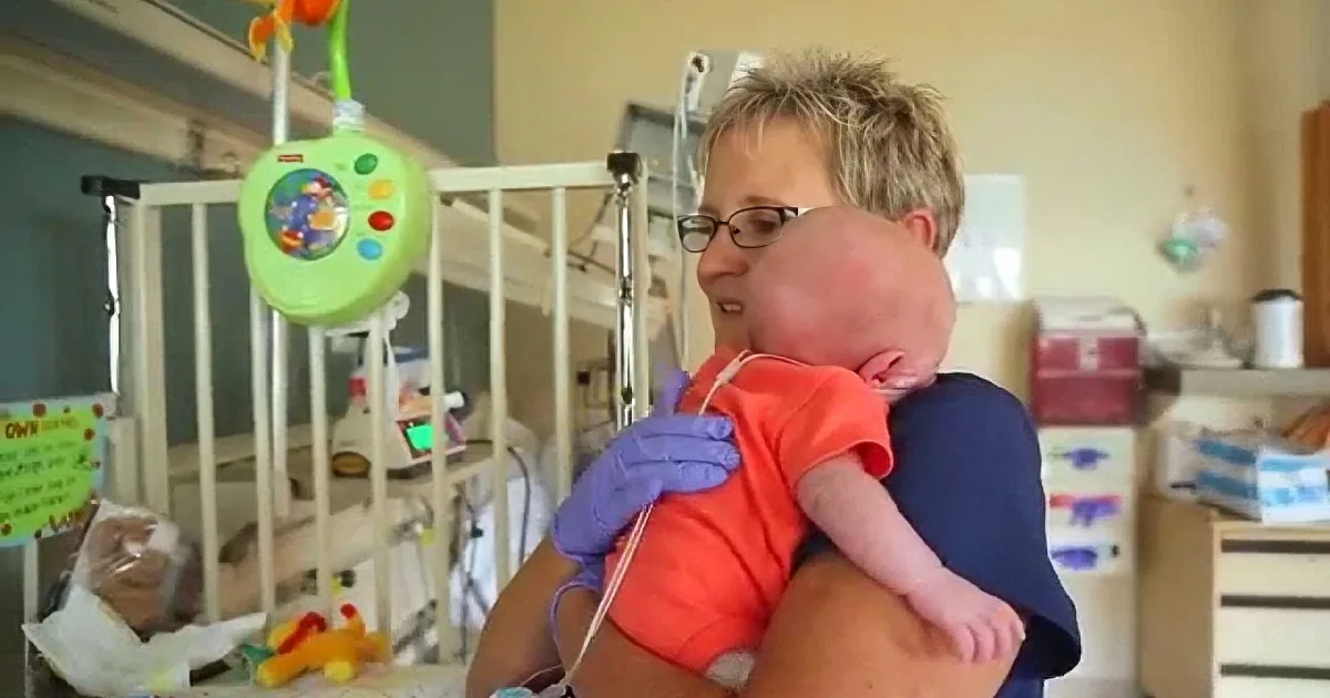 A woman holds a baby in a hospital setting, surrounded by medical equipment and toys.