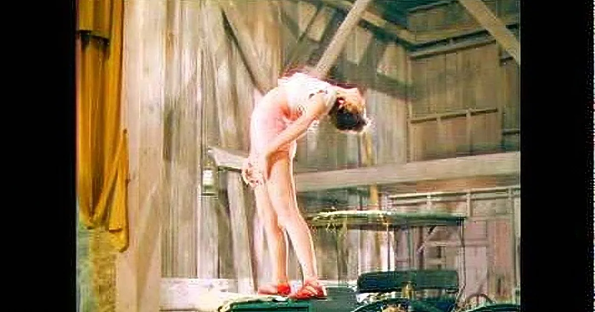 A nude person bends forward on a diving board in a wooden structure.