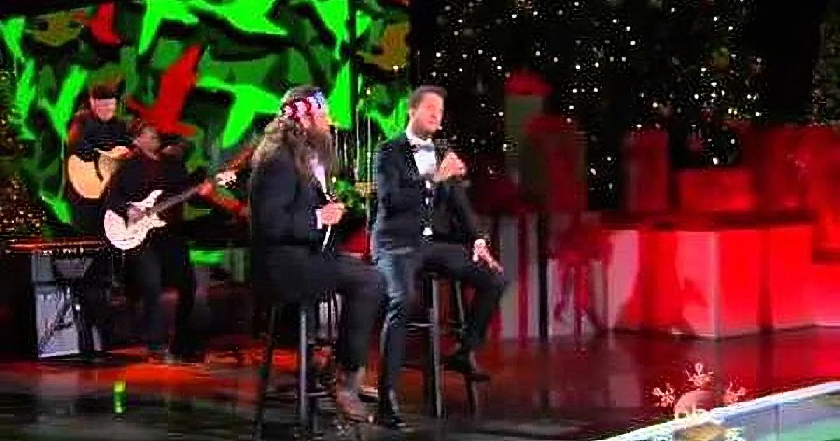 A man in a suit sits on a stool, singing into a microphone on a stage with a guitarist and a Christmas tree in the...