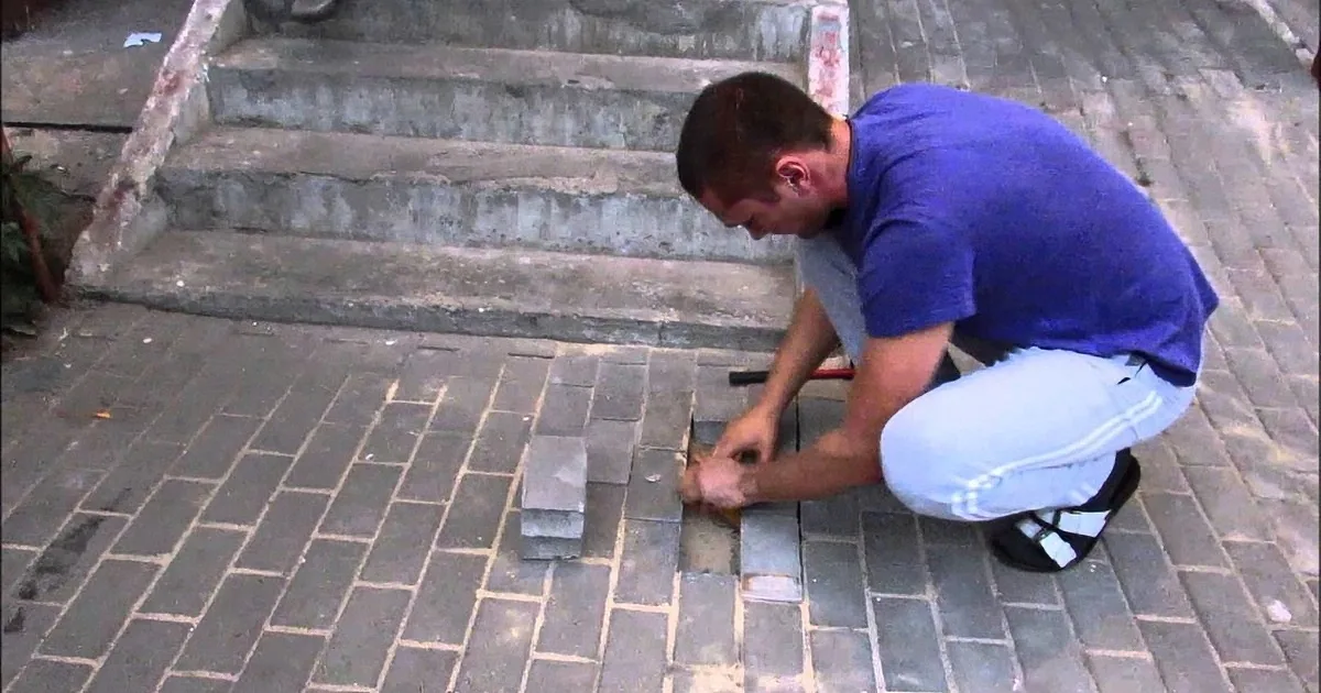 A man in a blue shirt and white pants is kneeling and working on a brick pavement.