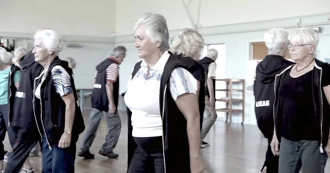 A group of older women in a dance studio, wearing black vests and white tops, practicing a synchronized movement.