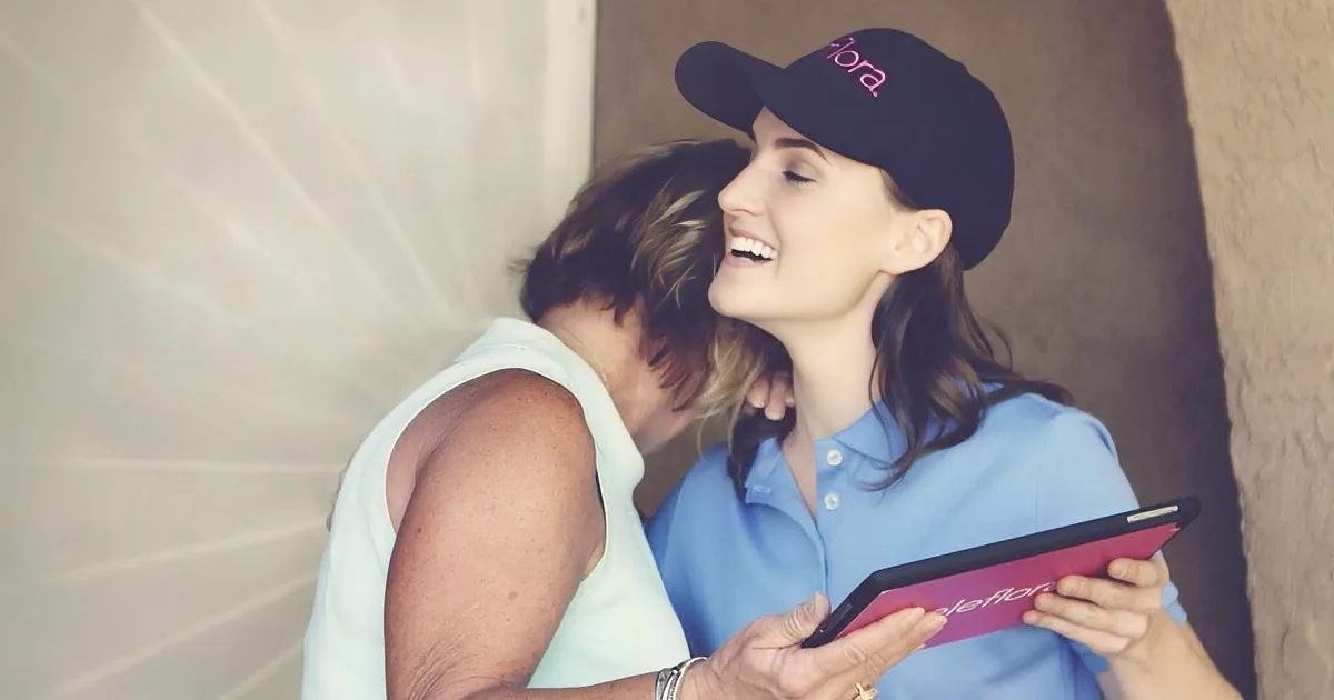 A woman in a blue shirt and black cap smiles while holding a tablet, interacting with another woman in a white top.