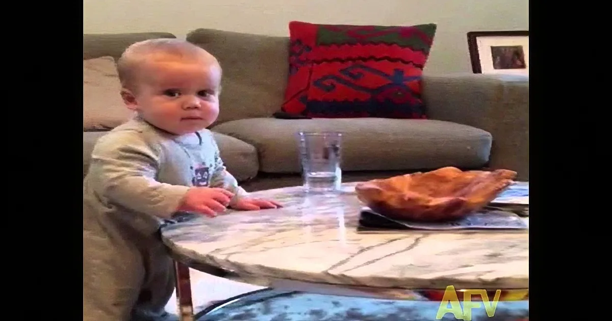 A baby stands near a table with a pizza and a glass of water, looking at something off-screen.