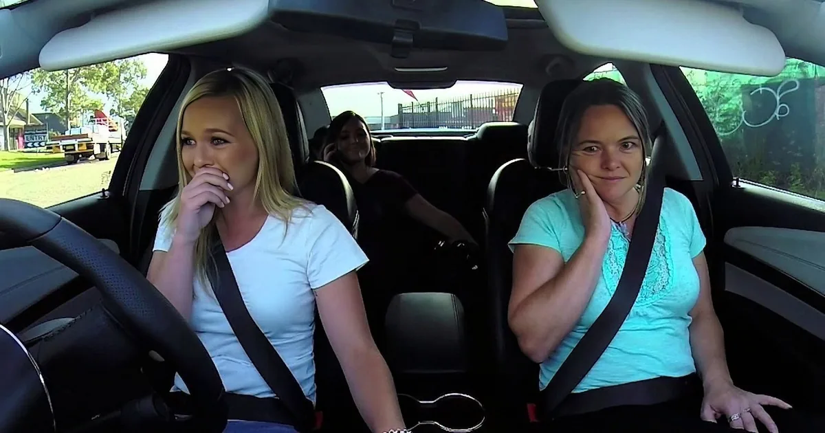 Two women in a car, one in the driver's seat, both wearing seatbelts, with a third person in the back seat.