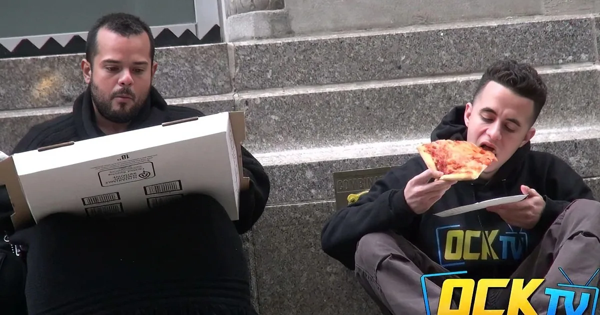 Two men sit on steps, one eating pizza, the other holding a pizza box, both wearing black jackets.