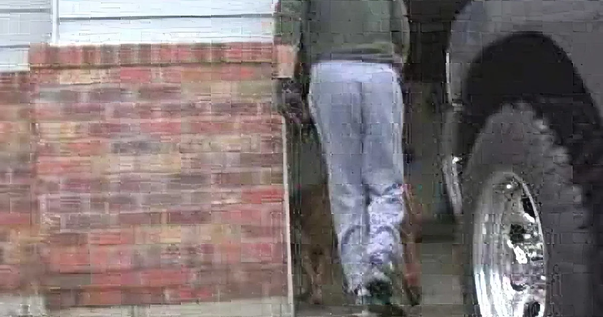 A person in a green jacket and gray pants walks past a brick wall and a vehicle.