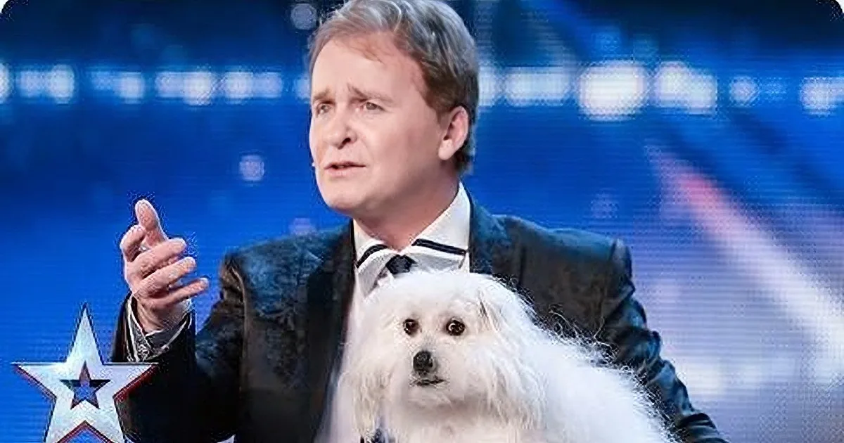 A man in a suit gestures while holding a fluffy white dog on a stage with a blue background.