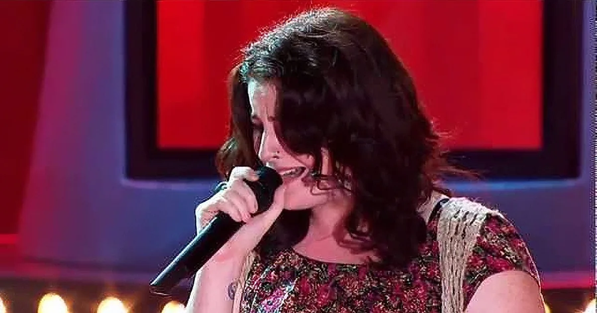 A woman with curly hair sings into a microphone, wearing a floral dress, with red and blue lighting in the background.