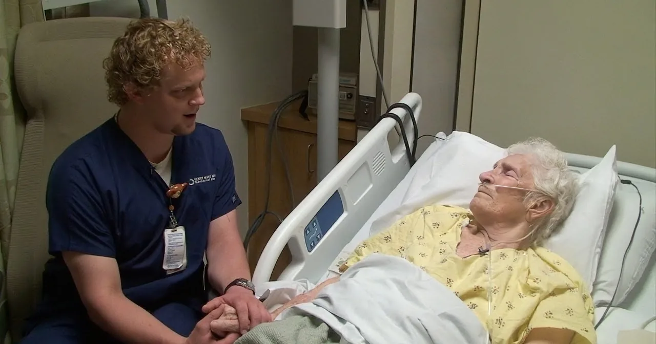 A nurse comforts an elderly patient in a hospital bed, holding hands.