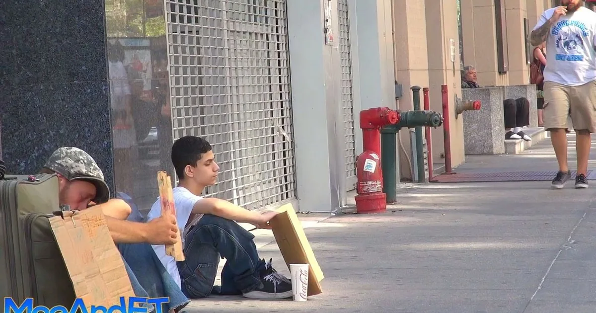 A man sits on the sidewalk holding a cardboard sign, with a Coca-Cola cup nearby.