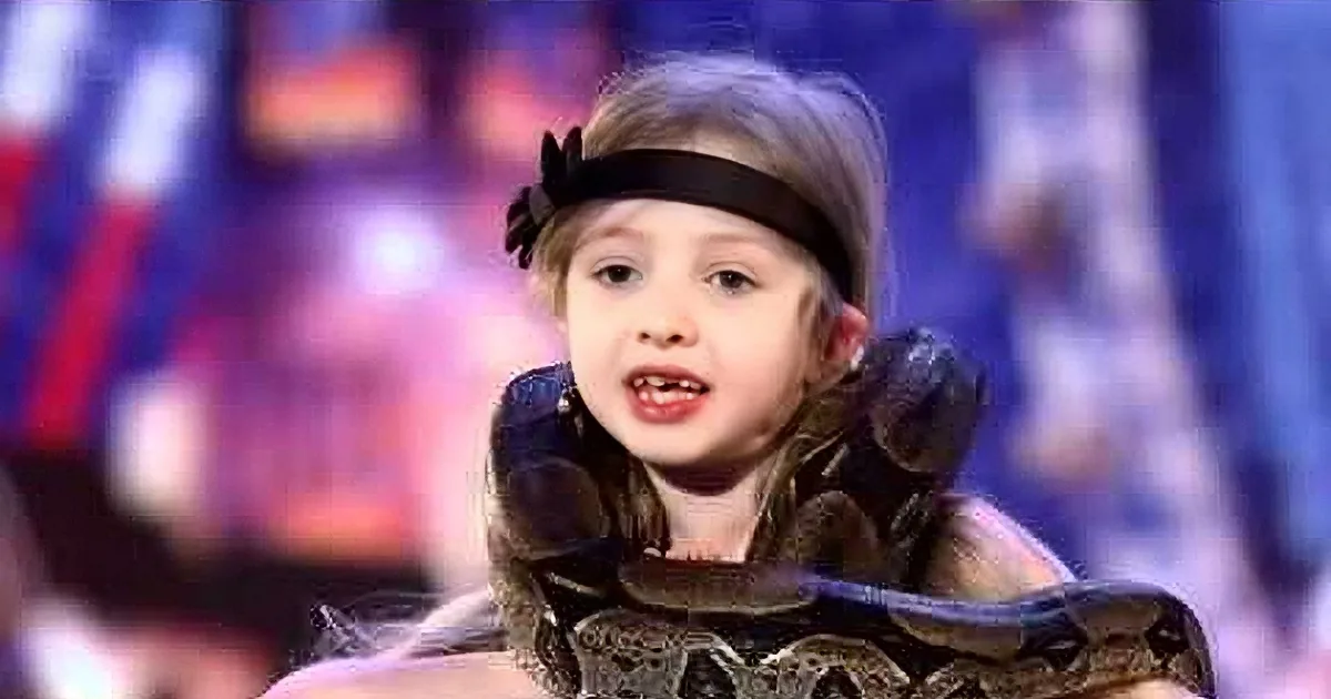 A young girl with a headband and snake around her neck, surrounded by blurred background.