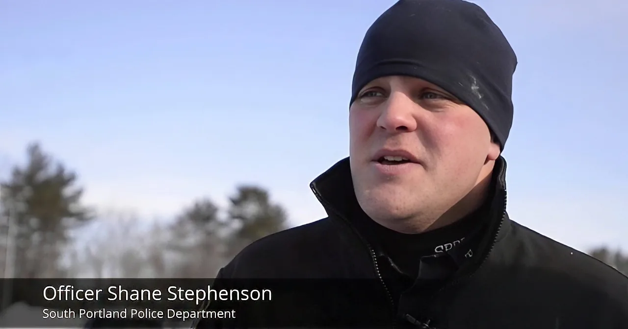 Officer Shane Stephenson from South Portland Police Department is speaking outdoors, wearing a black jacket and beanie.