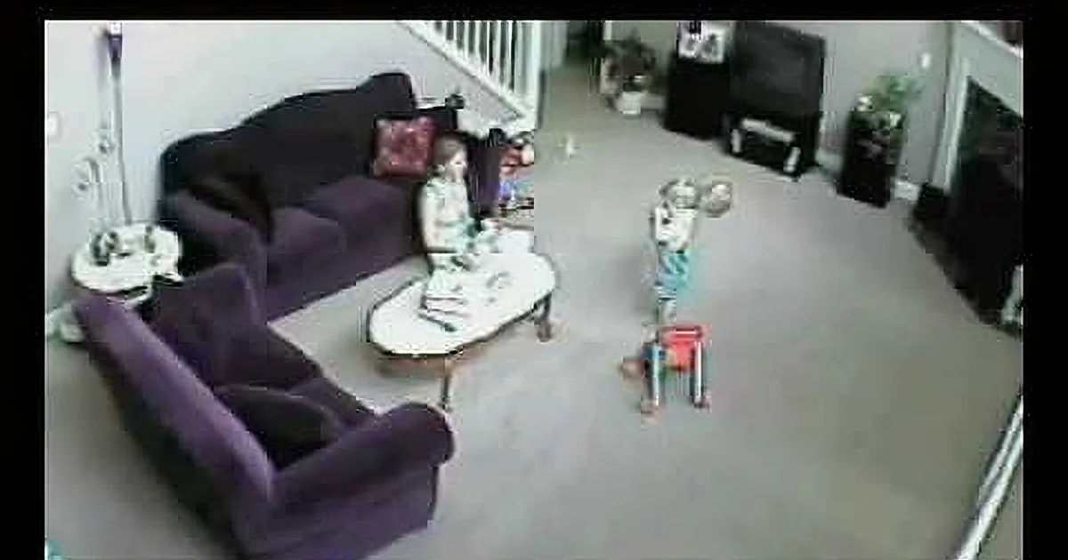 A child stands on a toy box in a living room with purple couches and a staircase in the background.
