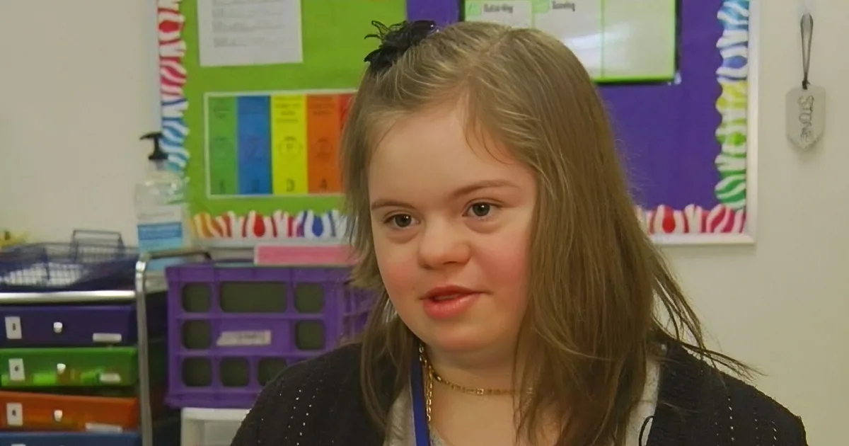 A young girl with long hair, wearing a black sweater and a blue lanyard, sits in a classroom with colorful posters and...