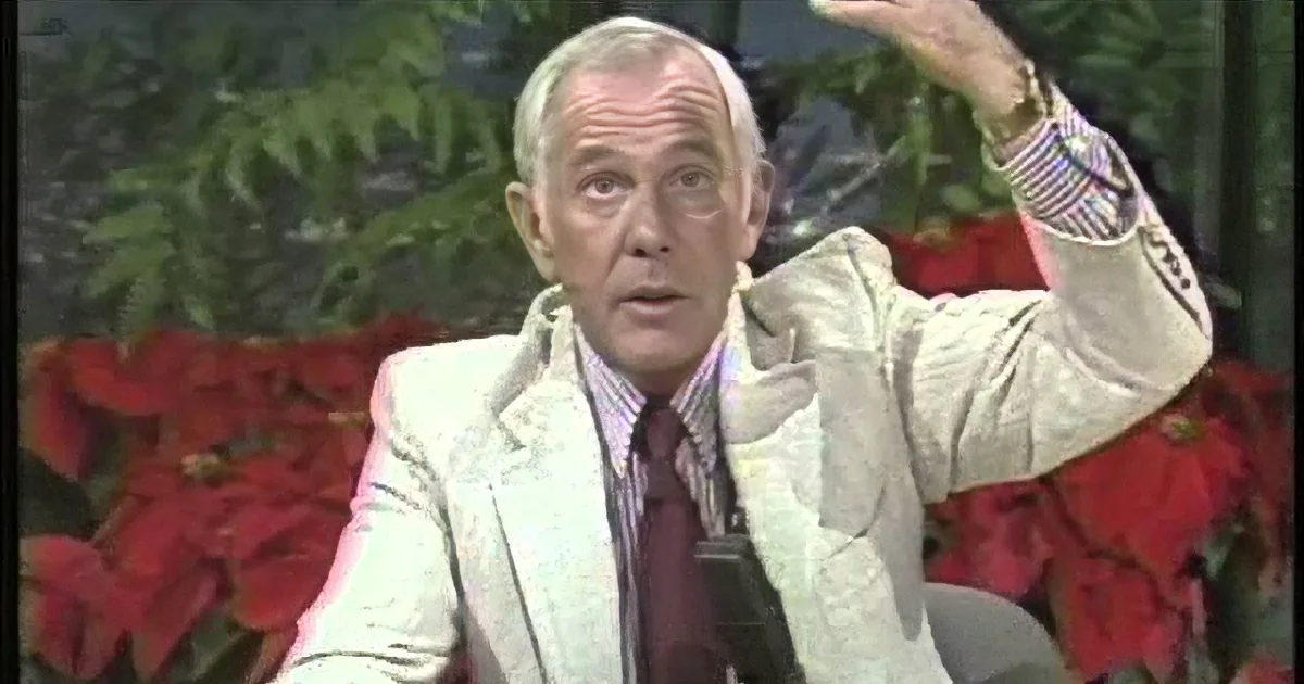 A man in a white suit gestures with his right hand, surrounded by red flowers and greenery.