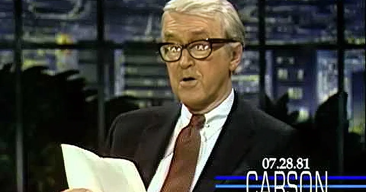A man in a suit and glasses holds a paper, appearing to speak on a news set.