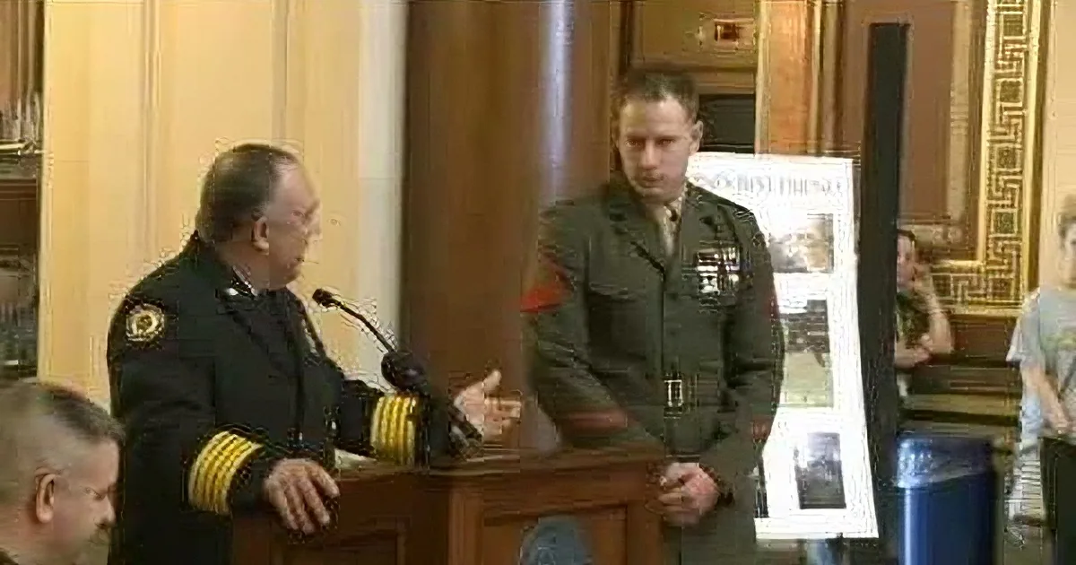 A man in a military uniform stands at a podium, addressing an audience, with another uniformed individual beside him.
