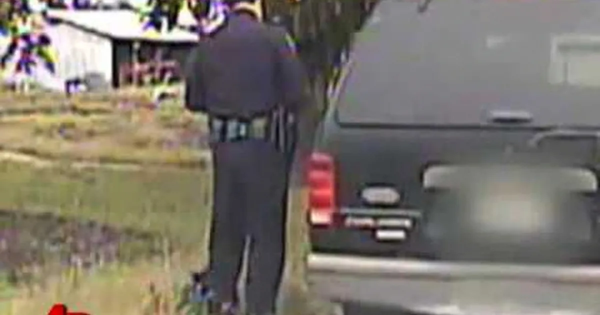 A man in a dark suit walks away from a black vehicle in a grassy area.