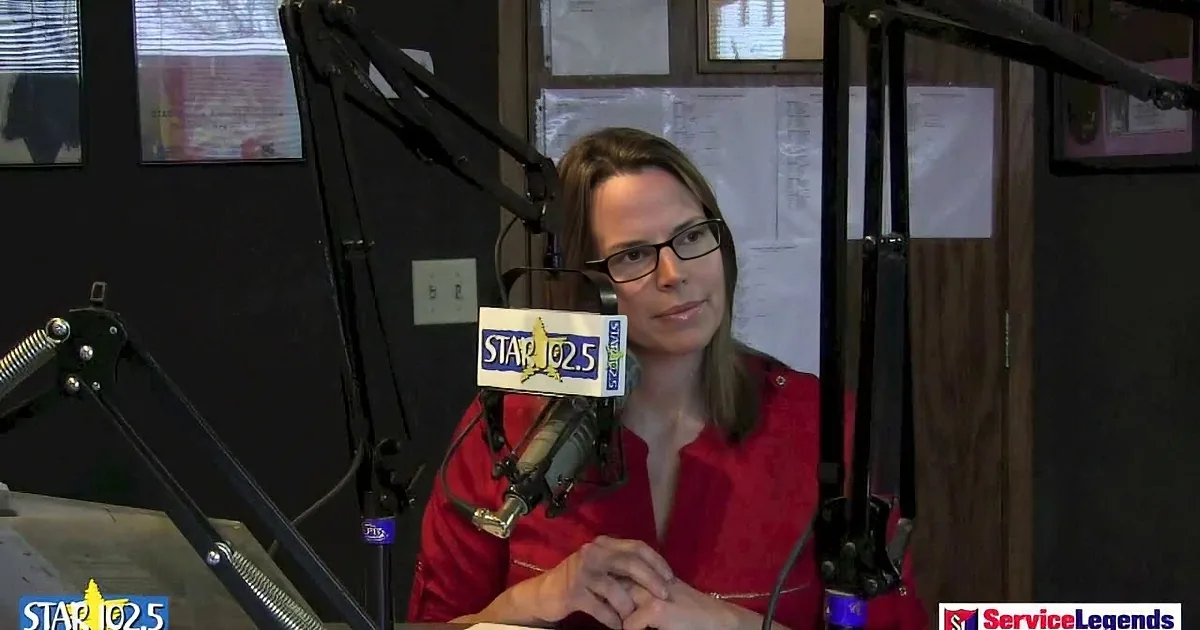 A woman in a red shirt sits at a radio station desk, speaking into a microphone.