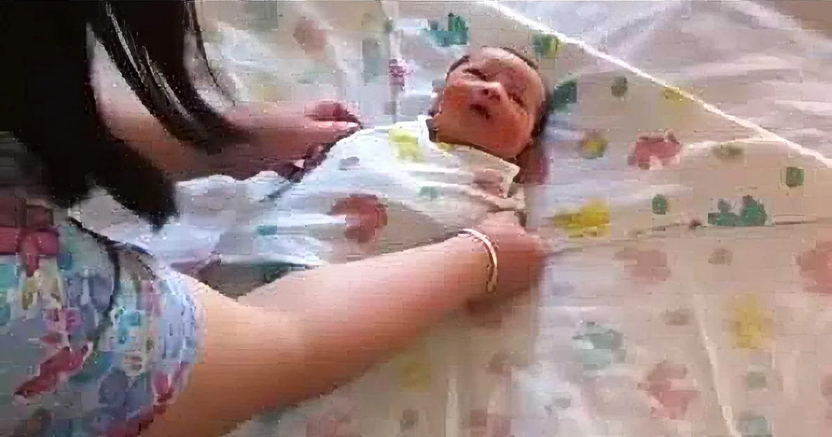A newborn baby is swaddled in a colorful blanket, lying on a patterned sheet, with a person adjusting the swaddle.