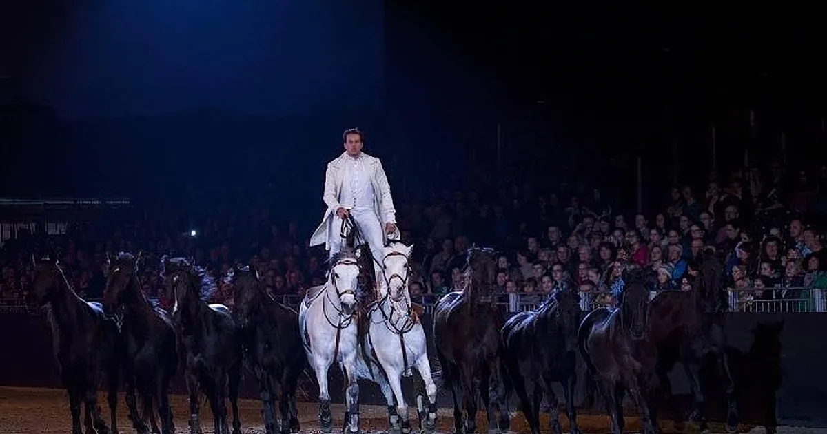 A man in white attire rides a white horse, leading a group of black horses in a performance.