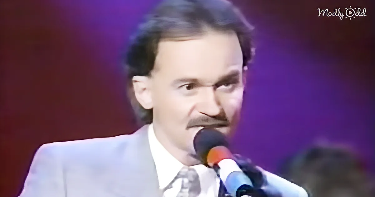 A man in a suit and tie sings into a microphone against a purple backdrop.