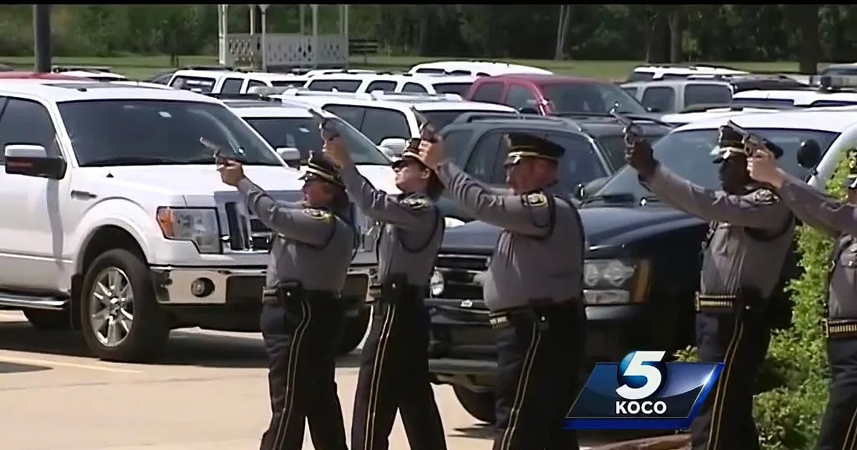 Police officers in uniform salute in front of a line of parked vehicles.