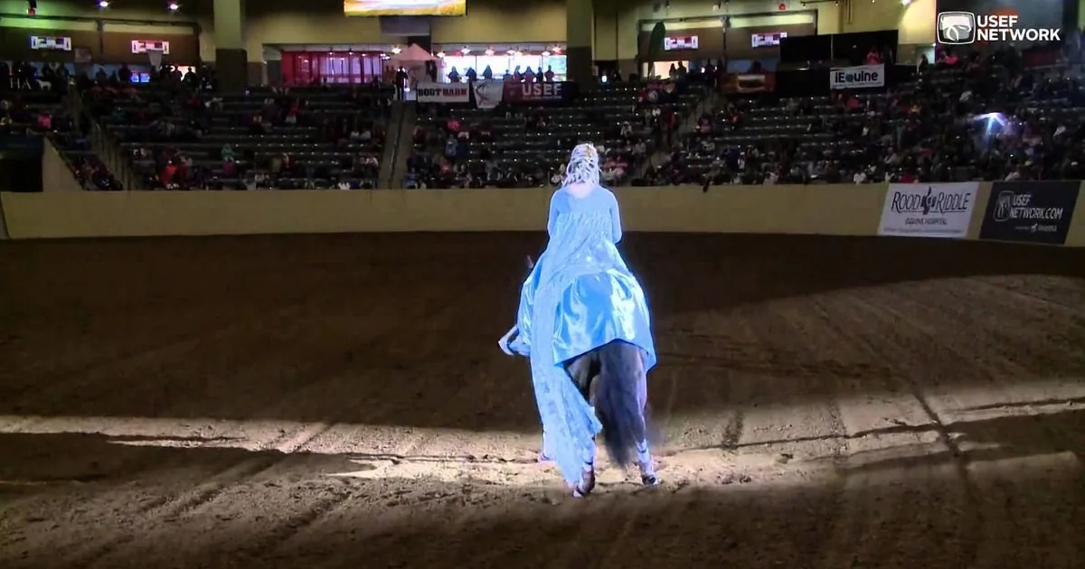 A person in a blue dress rides a horse in an indoor arena with spectators.