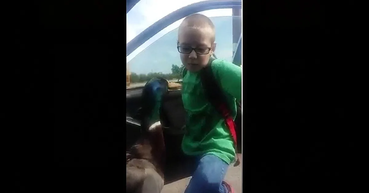 A child with glasses and a green shirt sits in a car, looking forward.