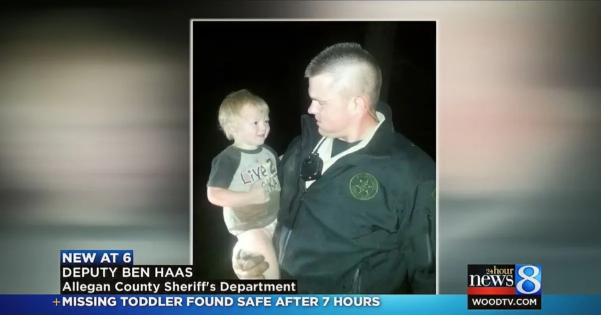 A deputy holds a smiling toddler, both in dark clothing, against a nighttime backdrop.