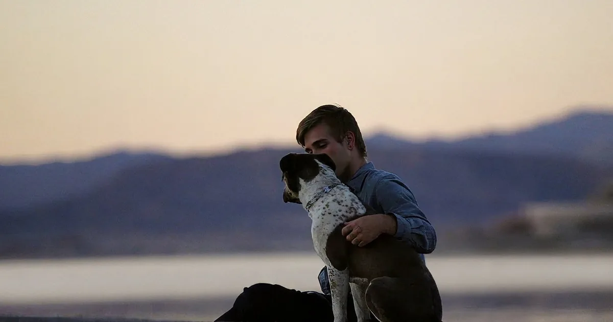 A man hugs a dog by a body of water with mountains in the background.