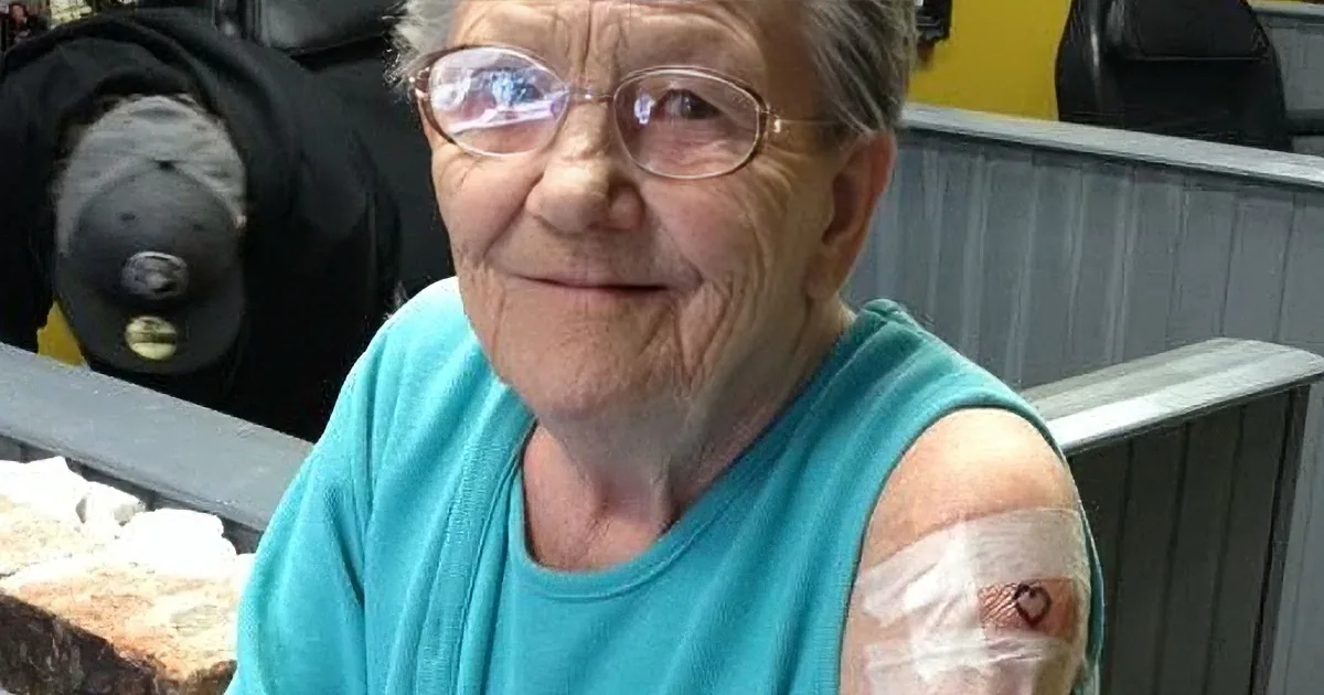 An elderly woman in a teal shirt shows a bandaged arm with a small heart tattoo after vaccination.