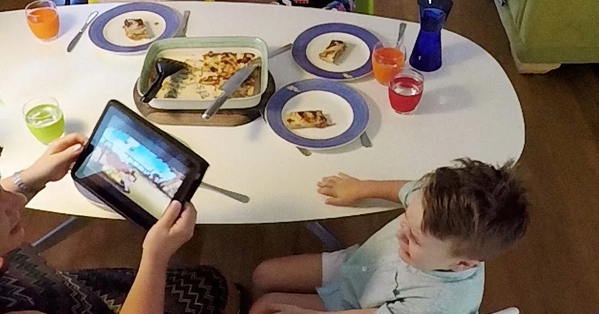 A child watches a tablet while another child sits nearby, with a table set for a meal featuring a casserole and drinks.