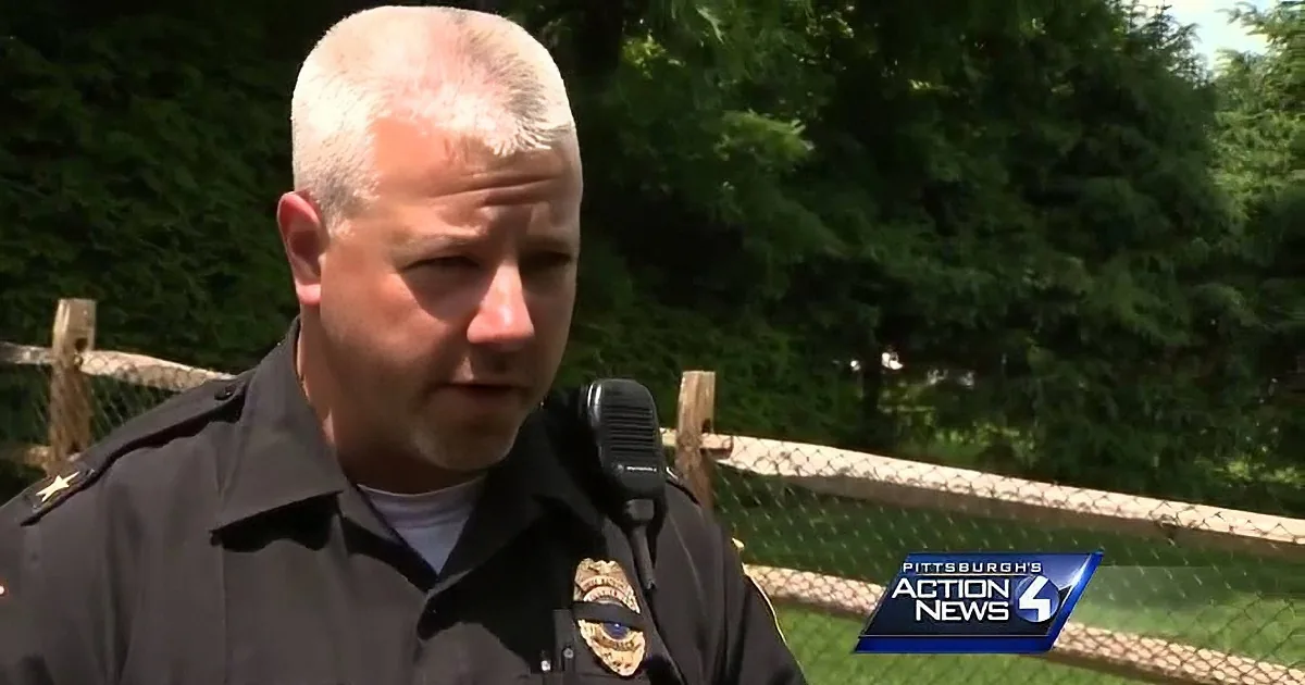 A police officer in uniform is speaking, with a background of trees and a fence.