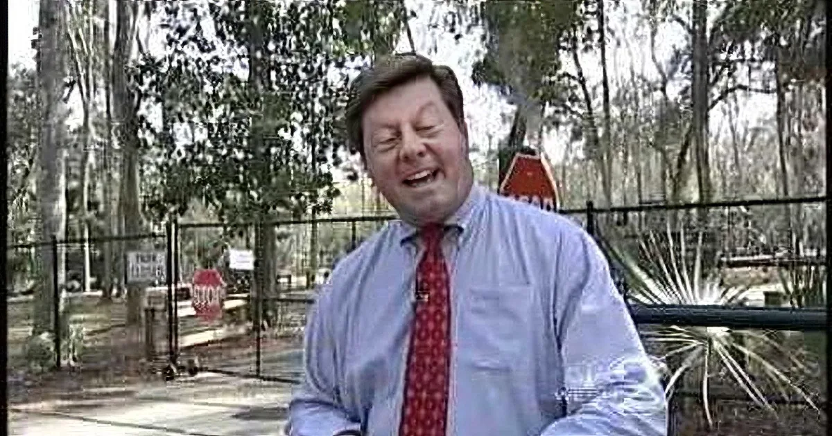 A man in a suit and tie is smiling outdoors, holding a glass, with trees and a fence in the background.