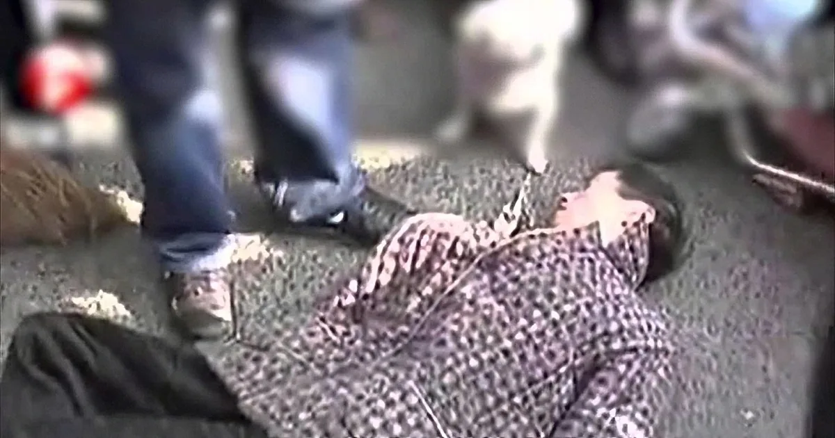 A person in black boots is standing over a body with a patterned cloth covering it.