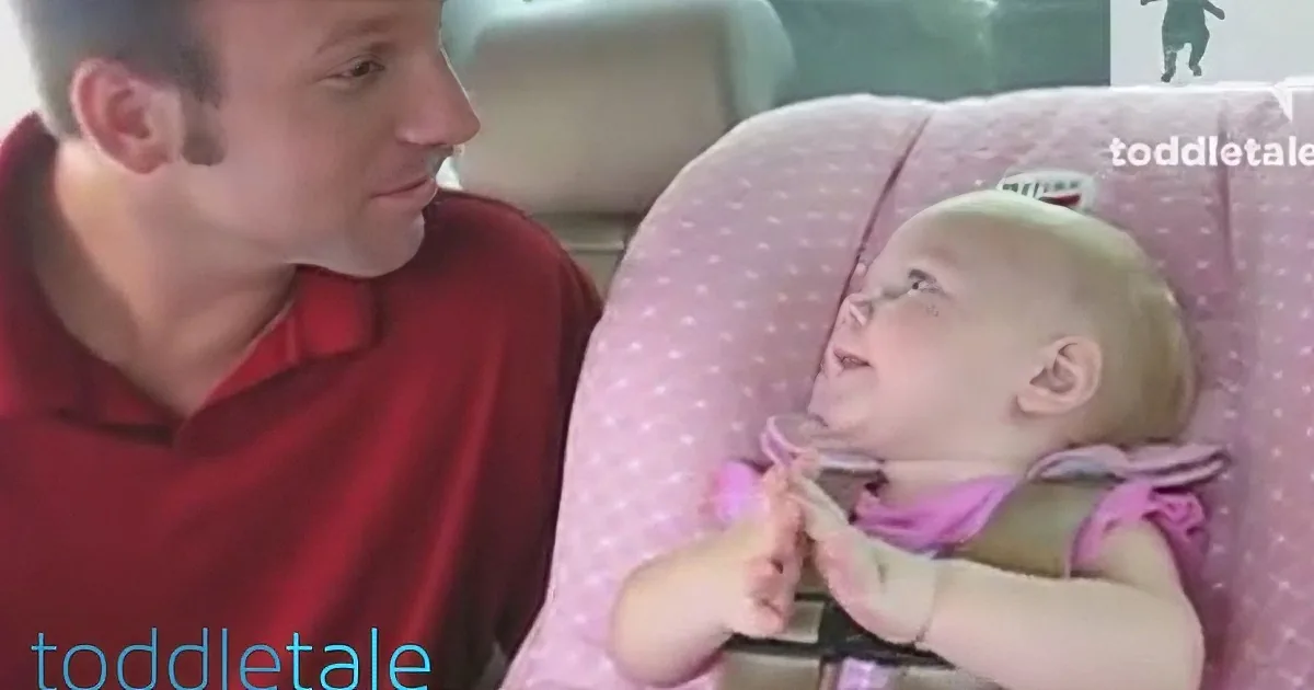 A man in a red shirt sits beside a baby in a pink high chair, both looking at each other.