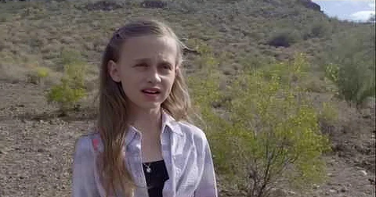 A young girl with long hair stands in a desert-like environment, wearing a white shirt over a black top.