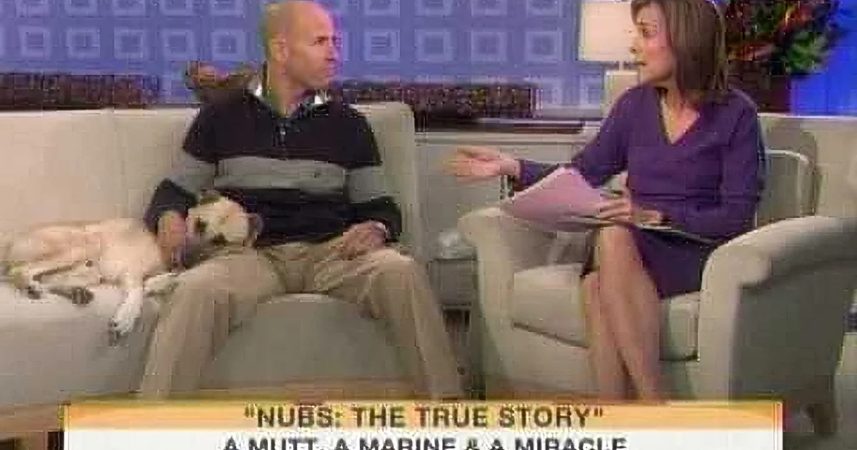 A man sits on a couch with a dog, talking to a woman on a TV show.