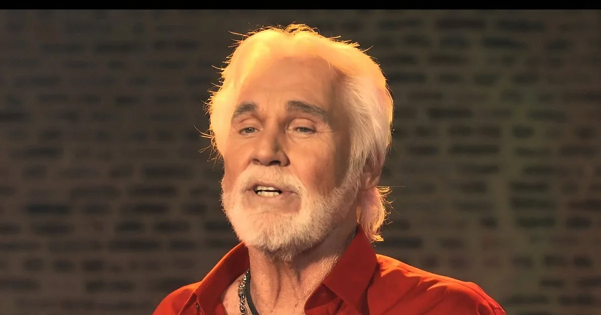 An older man with white hair and a beard, wearing a red shirt, stands against a textured background.
