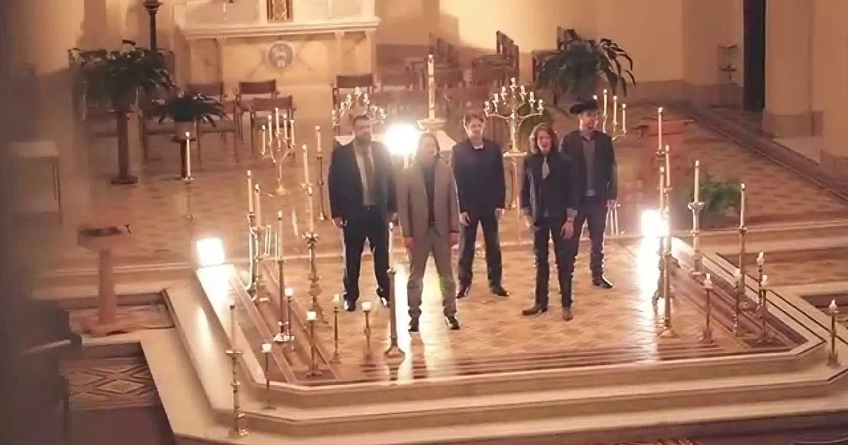 A group of six people in formal attire stands on a church altar surrounded by lit candles.