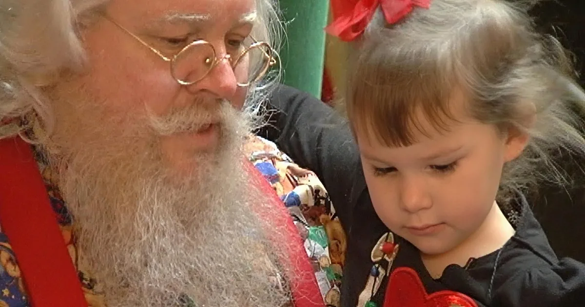 A bearded man in festive attire interacts with a young child wearing a red bow.