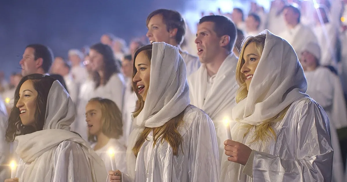 A group of people in white robes and head coverings hold candles, smiling and looking to the side.