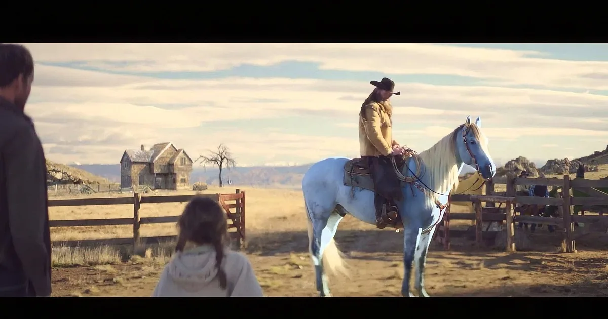 A man in a cowboy hat rides a white horse near a wooden fence, with a house and barren trees in the background.