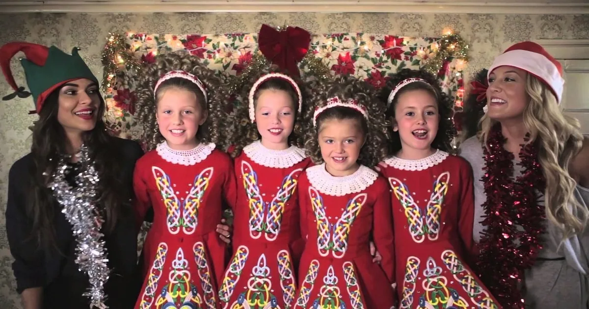 Five girls in red dresses with intricate designs, flanked by two women in festive attire, posing in front of a...