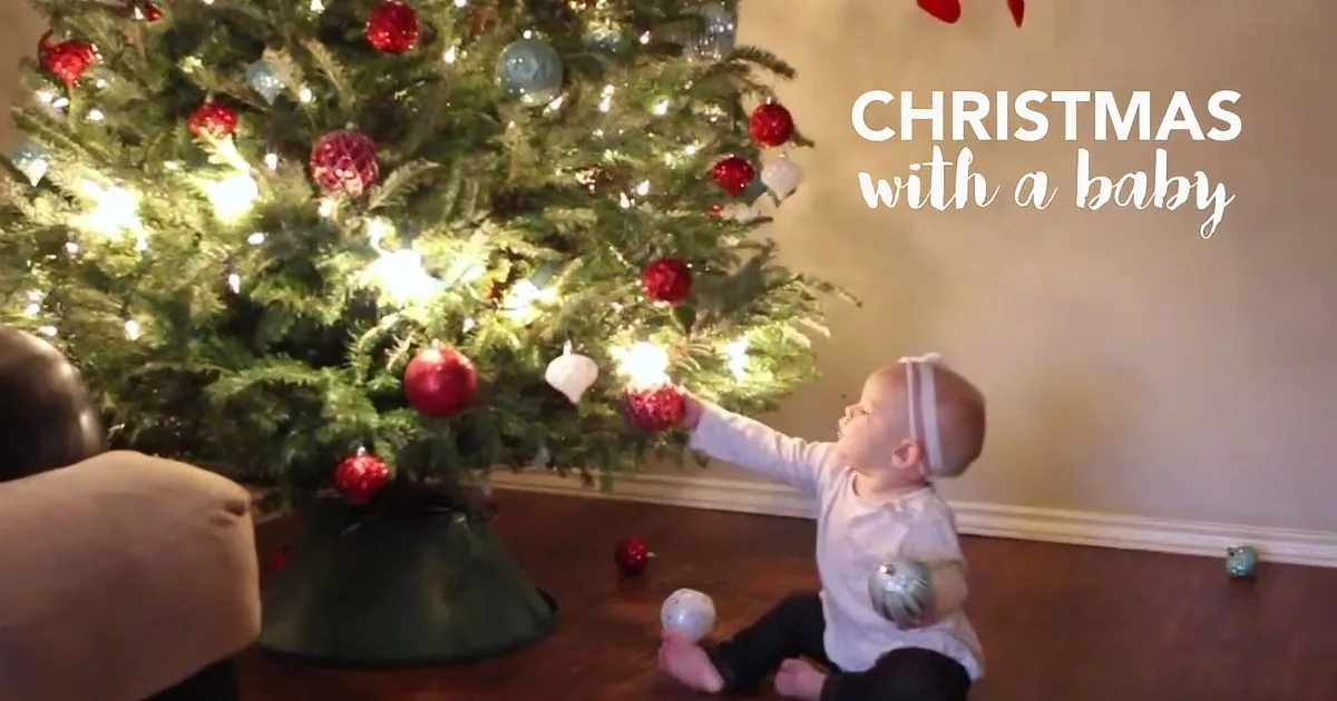 A baby decorates a Christmas tree with ornaments while an adult assists nearby.