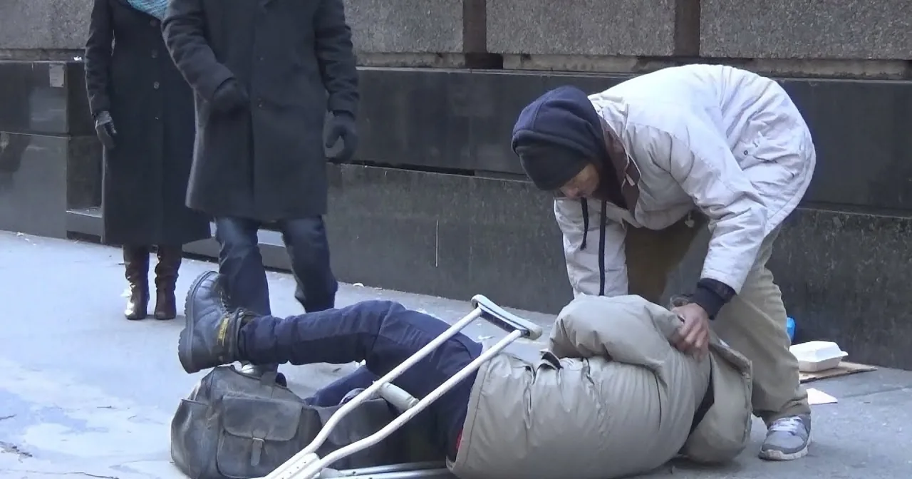 A person in a white coat helps another lying on the ground, with crutches nearby.
