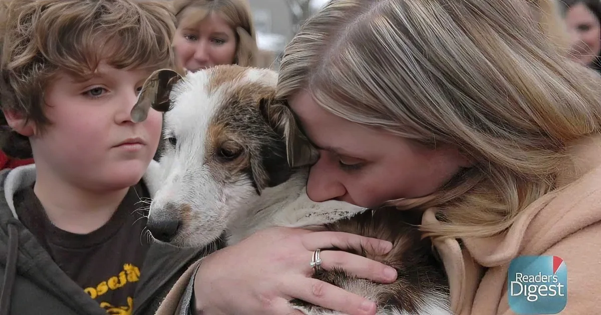 A woman hugs a dog while a boy watches, both smiling warmly.